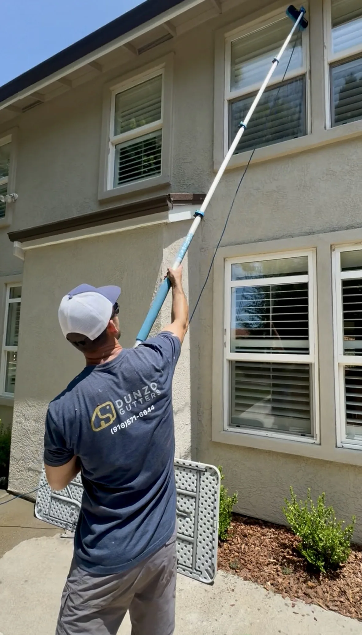 A Dunzo Gutters technician washing a second-story window with an extension pole and squeegee