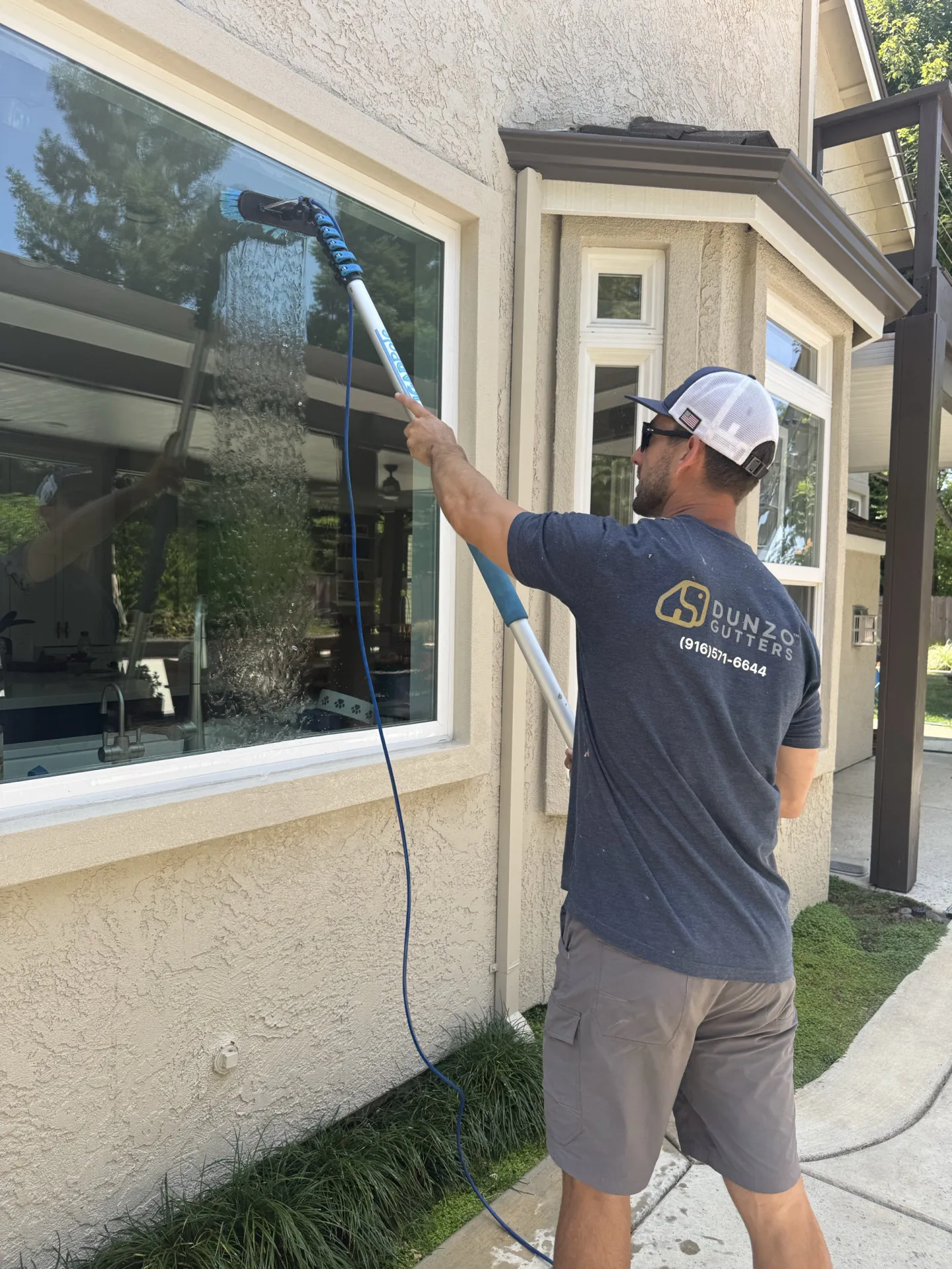 A Dunzo Gutters technician washing a window with an extension pole brush, water streaming down the glass